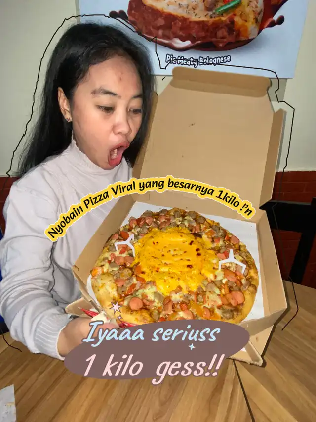 Seorang wanita dengan ekspresi terkejut memegang pizza besar 1kg dari Domino's Pizza, dengan tulisan 'Nyobain Pizza Viral yang besarnya 1kilo !'n' dan 'Iyaaa seriuss 1 kilo gess!!'.