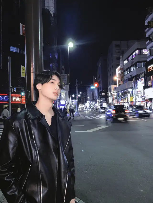 夜の新大久保の街中で、黒いレザージャケットを着た男性がカメラの方を少し見てポーズをとっている。背景には賑やかな夜の交差点が見える。