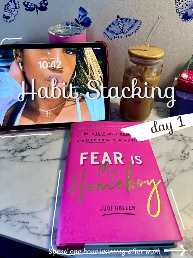 A flat lay shows a tablet displaying 'Habit Stacking' and a woman, alongside a drink and a pink book titled 'Fear Is My Homeboy' by Judi Holler. The image highlights 'day 1' of spending one hour learning after work, with butterfly decorations in the background.
