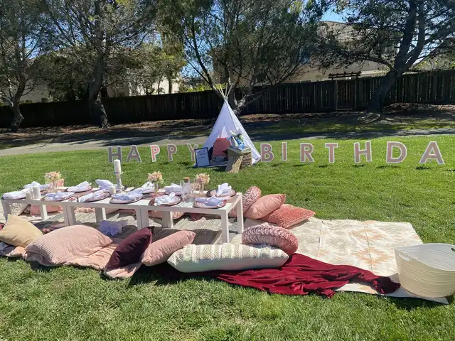 A wide shot of a luxury picnic birthday setup in a park, showcasing a low table, numerous cushions, and blankets on a patterned rug, with large 'HAPPY BIRTHDAY' letters and a white teepee tent.