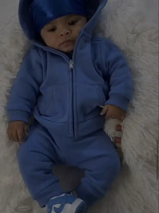 A baby in a blue hooded tracksuit and a blue head covering, lying on a white fluffy blanket, looking towards the camera.