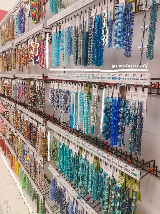 An aisle inside a craft store, showcasing numerous strands of colorful beads in various shades of blue, green, yellow, and orange, hanging on display racks. Text overlay says 'so many colors!'.