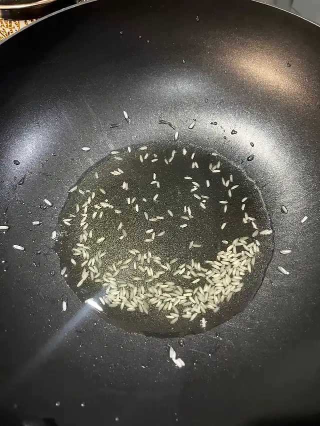 A black pan with a small amount of oil and a few grains of uncooked rice, illustrating the initial step of sautéing the rice.