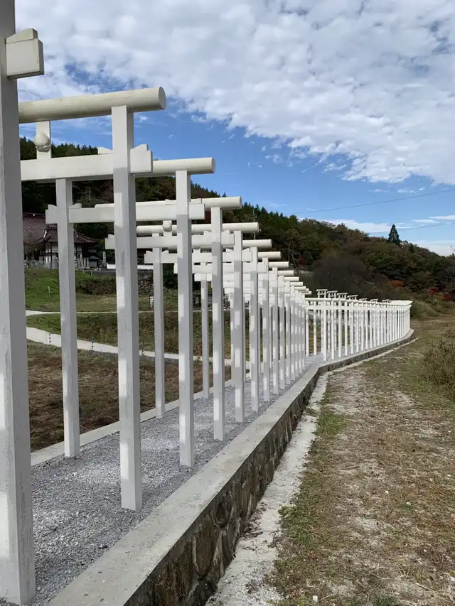 白い連鳥居が右にカーブしながら続く様子を側面から捉えた写真です。鳥居の横には砂利道と石積みの壁があり、遠くには山と雲のある青空が見えます。