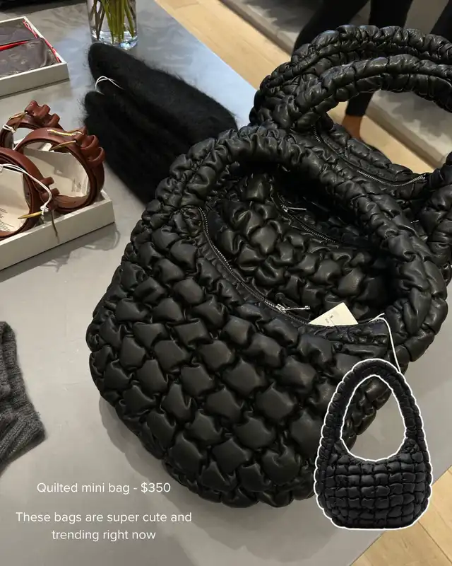 A close-up of a black quilted mini bag with a textured, puffy design, displayed on a table. The bag is described as a trending accessory, perfect for fall/winter outfits.