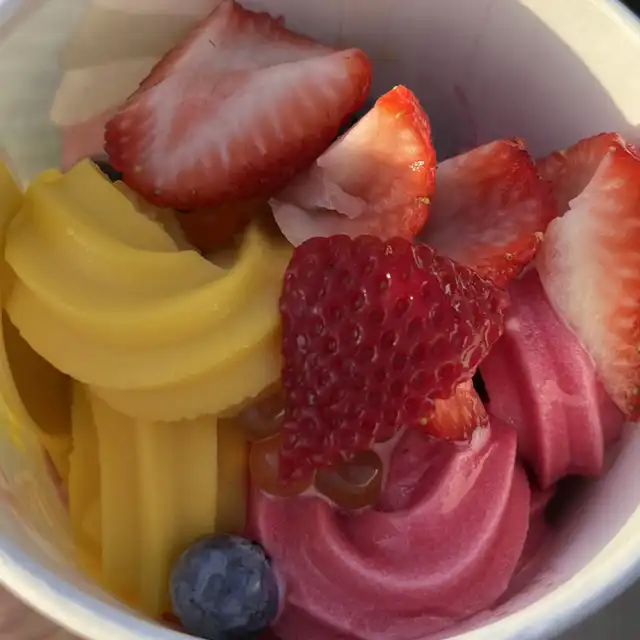 A white cup contains swirls of yellow mango and pink pomegranate frozen yogurt. It is topped with fresh sliced strawberries, a whole blueberry, and a sweet liquid drizzle.