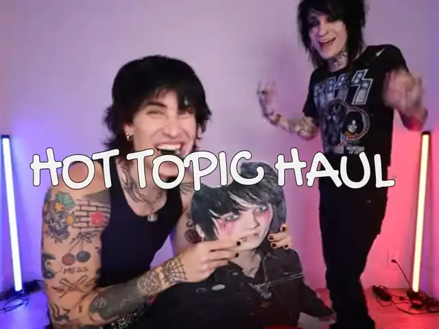 Two individuals with alternative style, one holding a cardboard cutout, are featured in a video thumbnail with the text "HOT TOPIC HAUL" overlayed. The background includes vertical LED light strips.