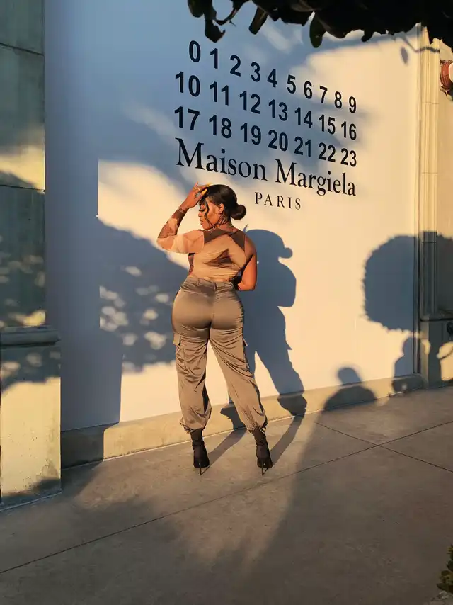 Ellejayy, a Los Angeles-based photographer, poses from behind in cargo pants and a sheer top in front of a white wall with "Maison Margiela Paris" text.