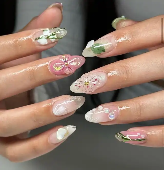 A close-up of hands displaying long, almond-shaped nails with delicate spring-themed designs. Features include 3D flowers, green leaf accents, pearls, and clear, pink, and white bases, creating a fresh and elegant look.