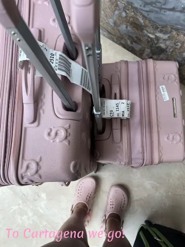 A top-down view of two pink suitcases with luggage tags showing destinations like CTG and MIA, placed on a tiled floor. A person's feet wearing pink sandals are visible between the suitcases, with text overlay "Cartagena we go!".