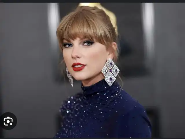 Taylor Swift is shown in a close-up, wearing a dark blue sequined high-neck top and large diamond-shaped earrings with purple details. She has blonde hair with bangs and bold red lipstick.