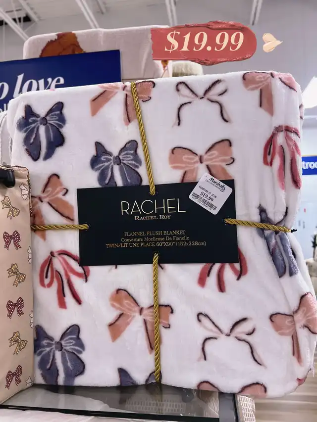 A folded white flannel plush blanket adorned with a pattern of pink, blue, and peach bows, secured with a gold cord. A price of $19.99 is displayed.