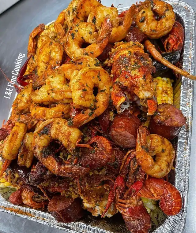 A large foil tray generously filled with a vibrant seafood boil, featuring seasoned shrimp, crawfish, corn on the cob, and sliced sausage. 'L&E Fine Cooking' is visible.