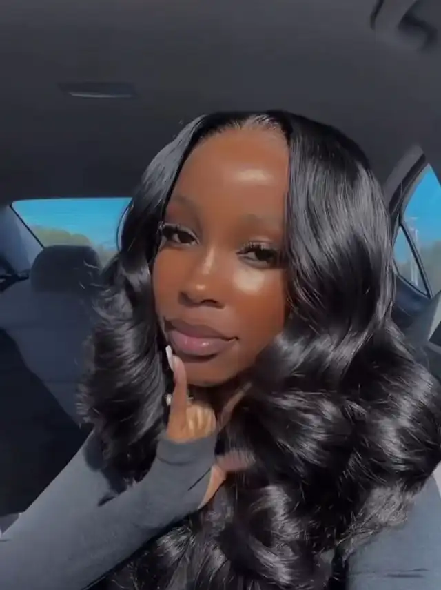 A woman with long, wavy black hair styled with a middle part, posing in a car. Her hair has a glossy finish and soft curls.
