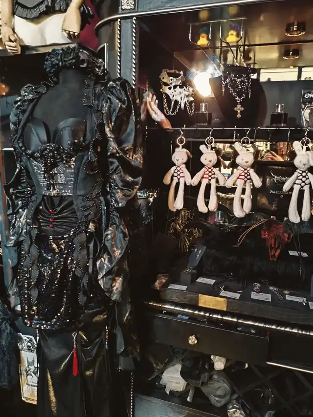 A black gothic-style dress on a mannequin is displayed alongside various accessories, including necklaces and bunny-shaped keychains, in a dark-themed shop setting.