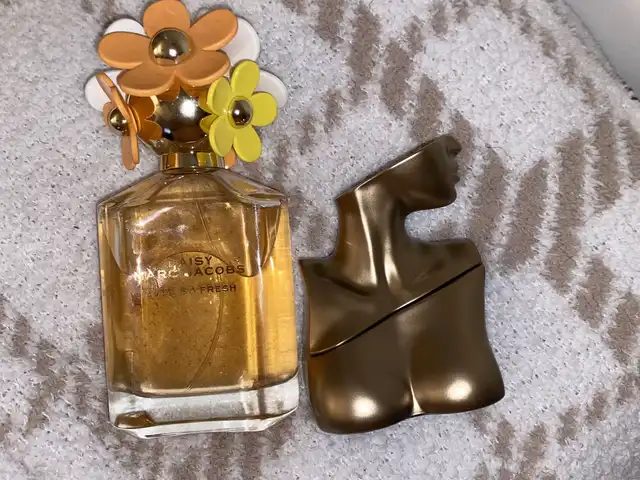 Two distinct fragrance bottles are shown on a textured surface: one is a clear bottle with a gold liquid and a floral cap, and the other is a gold, bust-shaped container.