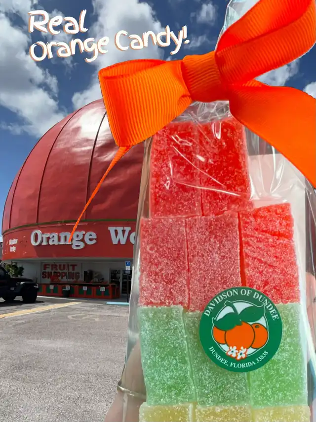 A clear bag of red and green fruit candies, labeled "Davidson of Dundee," is held in front of the Orange World building. An orange ribbon adorns the bag, and the text "Real Orange Candy!" is overlaid.