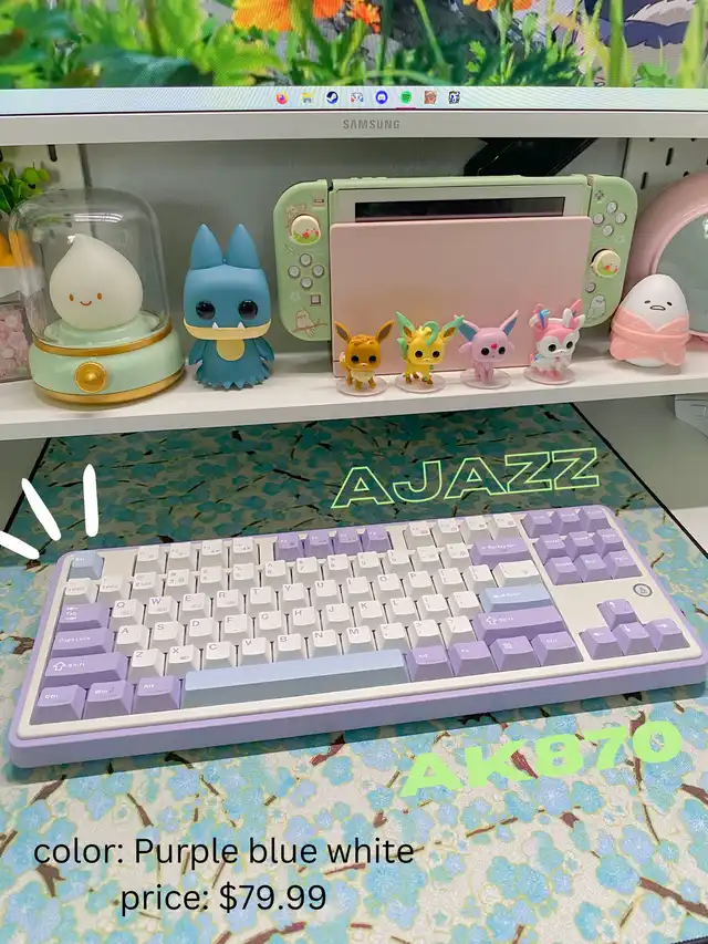 A close-up of a mechanical keyboard with purple, blue, and white keys, identified as the Ajazz AK870, with text indicating its color and price. The background shows a desk setup with a monitor and figurines.