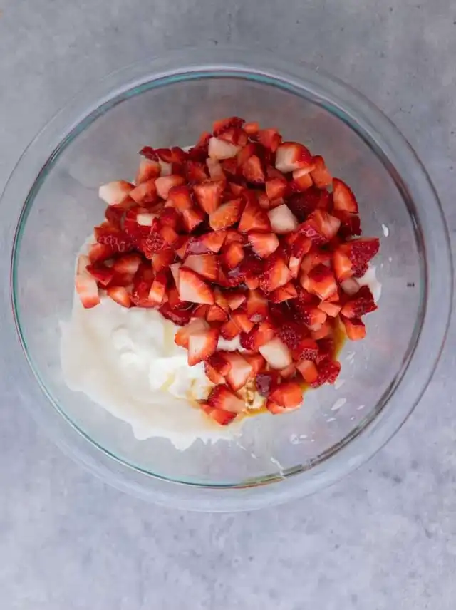 A clear glass bowl containing plain Greek yogurt, honey, vanilla extract, and a pile of diced fresh strawberries, ready to be mixed for the yogurt clusters.