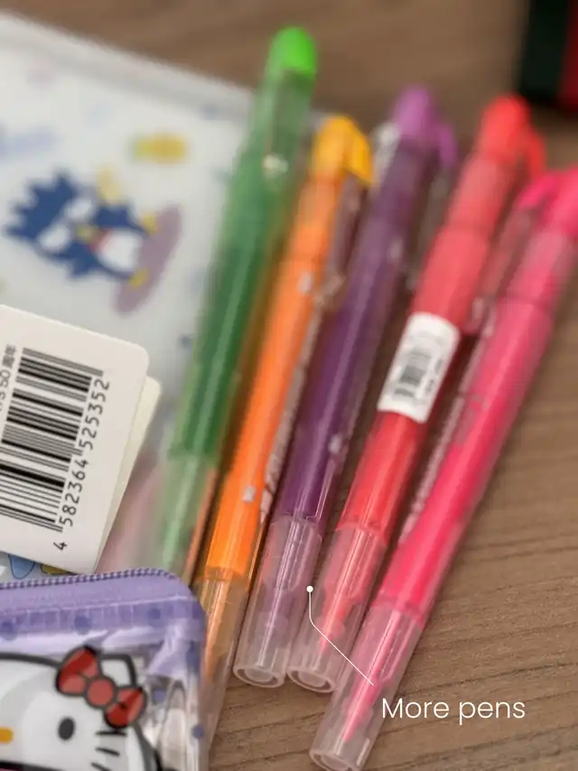 A close-up of five colorful pens in green, orange, purple, red, and pink, with a small purple Hello Kitty pouch partially visible. The text "More pens" is overlaid on the image.