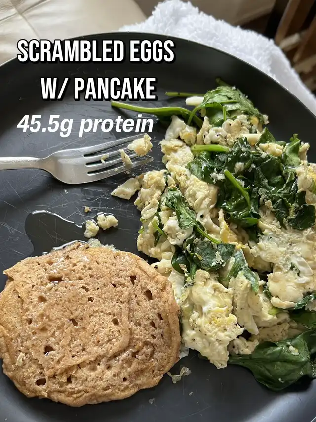A black plate holds scrambled eggs mixed with spinach and a round pancake, with a fork beside them. Text overlay reads "SCRAMBLED EGGS W/ PANCAKE 45.5g protein," showcasing a sweet and savory high-protein meal.