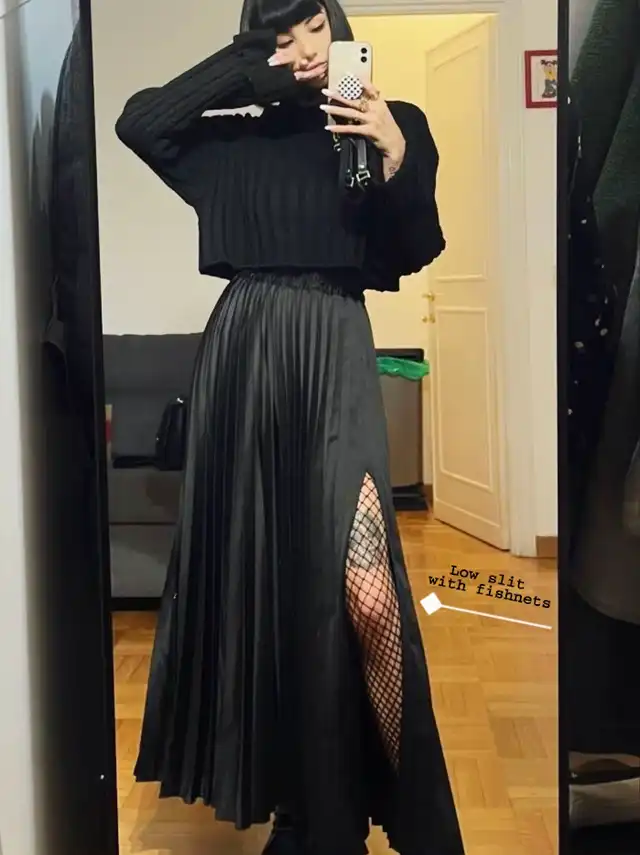 A person in a modest alt outfit featuring a black cropped long-sleeve top and a long black pleated skirt with a high slit, revealing fishnet stockings underneath. Text overlay points to "Low slit with fishnets."