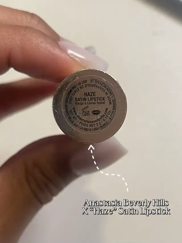 A hand holding the bottom of a lipstick tube, labeled "HAZE SATIN LIPSTICK," with an overlay identifying it as a specific satin lipstick.